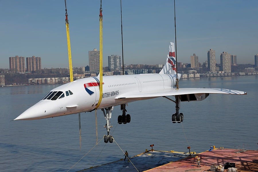 A retired British Airways Concorde is suspended above water by yellow cables and cranes, with a city skyline and river in the background.