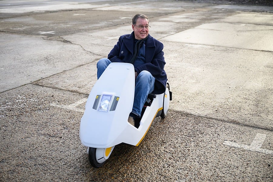 A person in a dark coat and jeans rides a small, white, three-wheeled electric vehicle on an empty, paved outdoor surface.