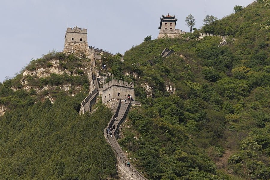 The Great Wall of China winds over a green, tree-covered hillside with watchtowers along its path. People are walking on the wall, and the sky is clear.