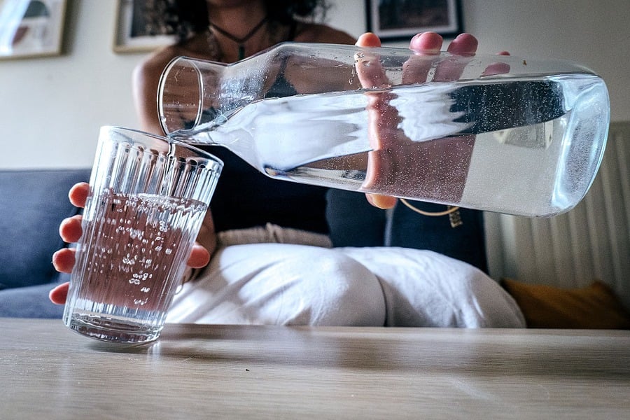 A person pours water from a clear glass pitcher into a ribbed glass tumbler, sitting at a table with a blurred background of a living room.