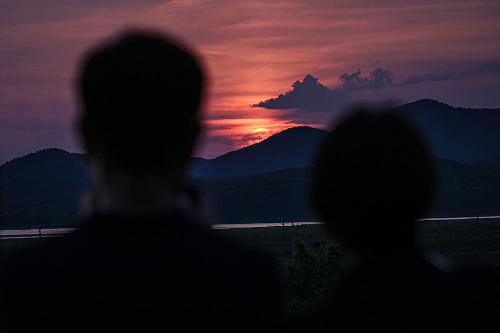 Two silhouetted figures watch a vivid sunset over distant mountains, with the sky painted in shades of pink, orange, and purple.