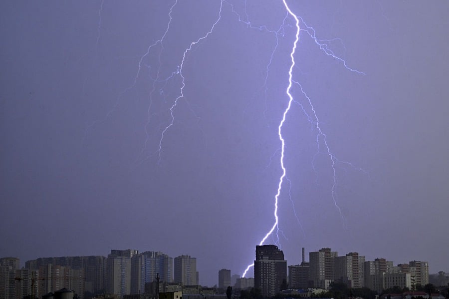 A bright bolt of lightning strikes above a city skyline at night, illuminating high-rise buildings under a dark, cloudy sky.