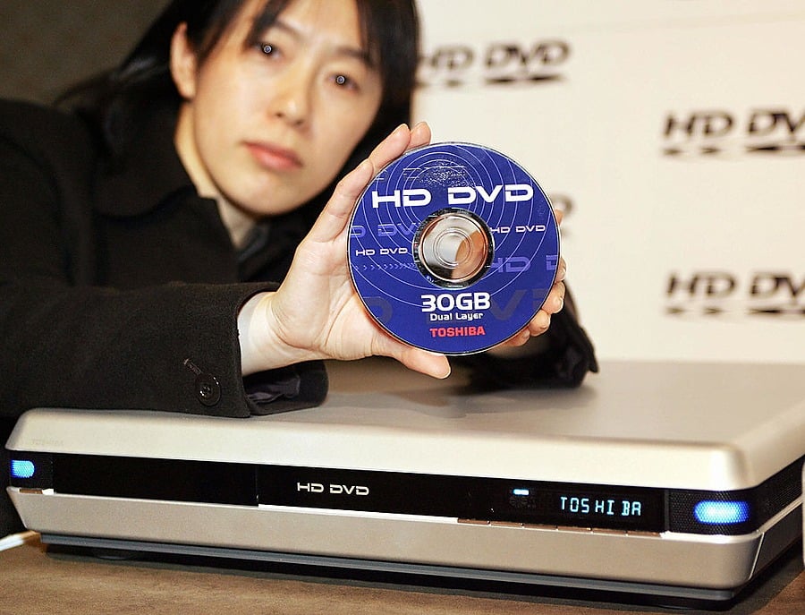 A person holds up a 30GB HD DVD disc in front of an HD DVD player. The player displays the brand “Toshiba” on its screen.