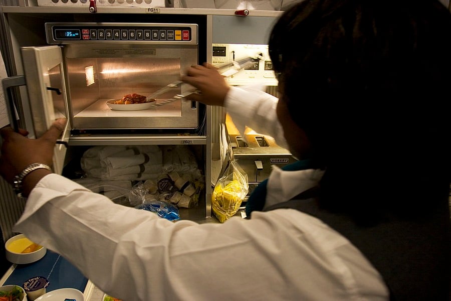 A person in a white shirt heats a plate of food in a microwave, likely inside an airplane galley, with various supplies and food items visible on the counter nearby.