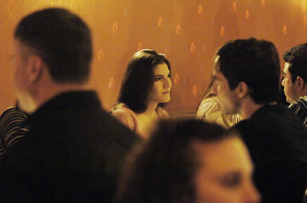 A woman with long brown hair sits at a dimly lit restaurant, looking thoughtfully at someone across the table. Several blurred people are in the foreground, creating a cozy, intimate atmosphere.