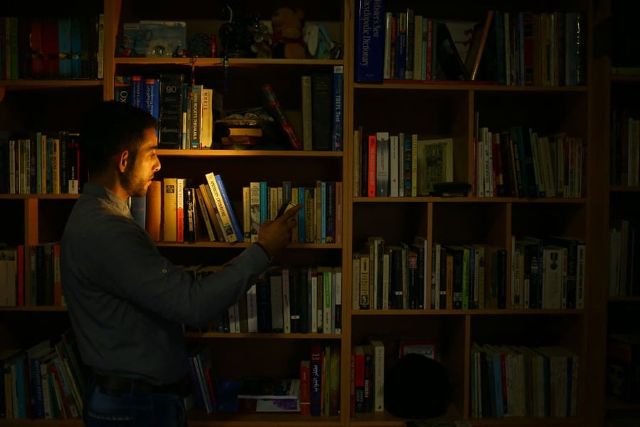 A man stands in front of a bookshelf, illuminated by a beam of light, looking at a phone in his hand. The room is otherwise dimly lit with many books visible on the shelves.