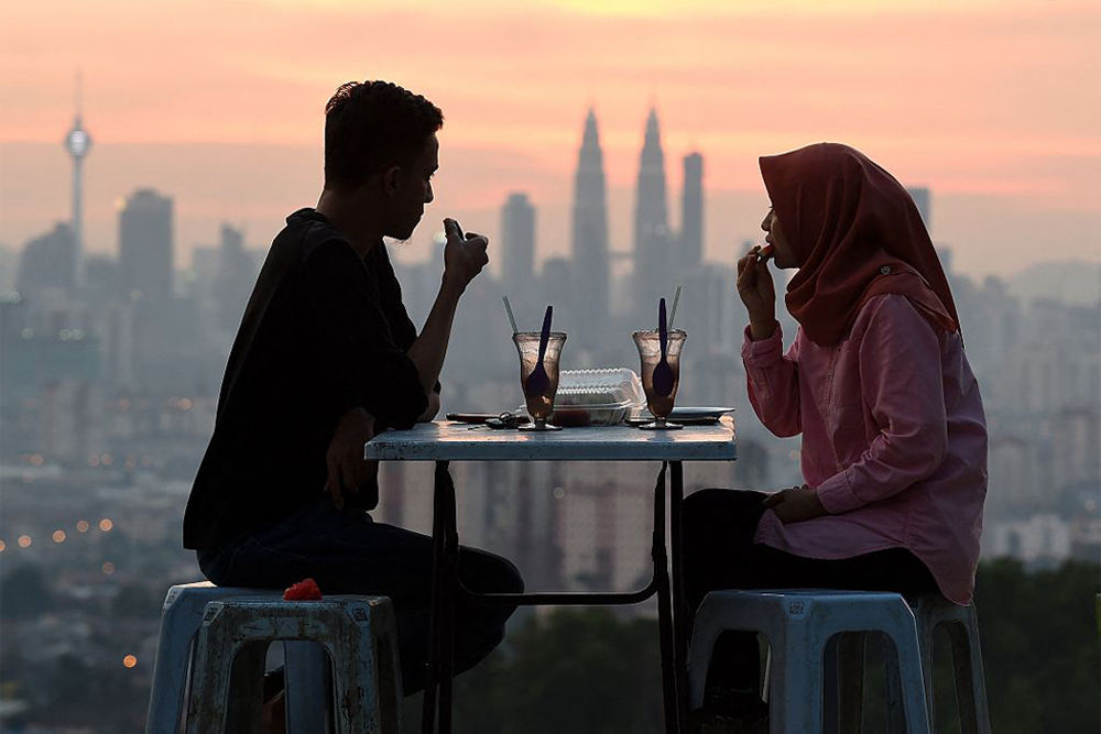Two people sit at a table outdoors, enjoying drinks and snacks at sunset with a city skyline, including the Petronas Twin Towers, visible in the background.
