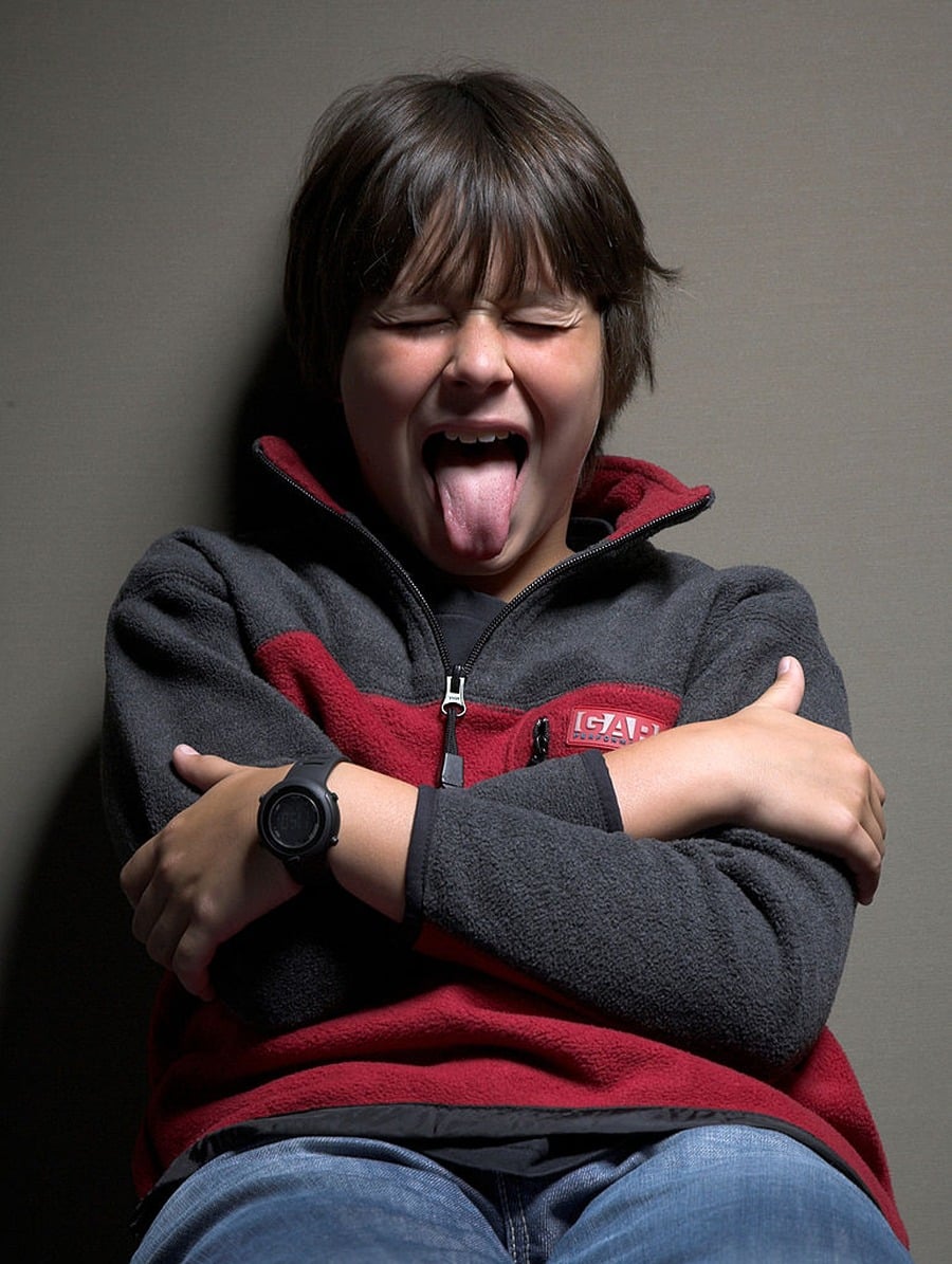 A boy in a red and gray fleece jacket sits against a wall, arms crossed, eyes closed, and sticking his tongue out playfully.