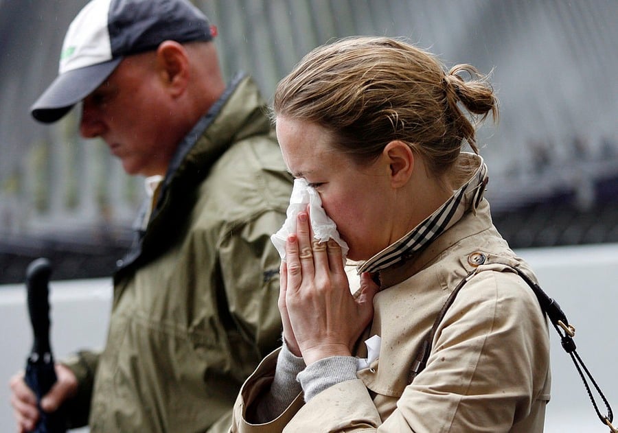 A woman in a beige coat wipes her face with tissues while a man in a green jacket and cap walks beside her outdoors.