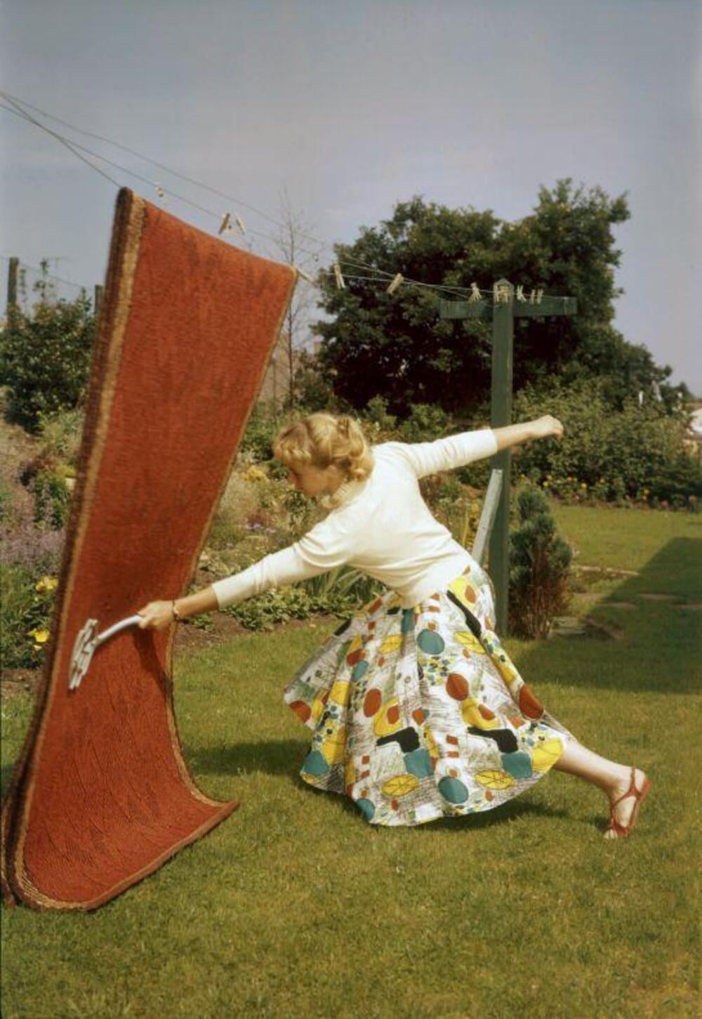 A woman in a colorful skirt beats a large red rug outdoors on grass, holding a rug beater with one hand, mid-swing, with gardens, a clothesline, and trees in the background.
