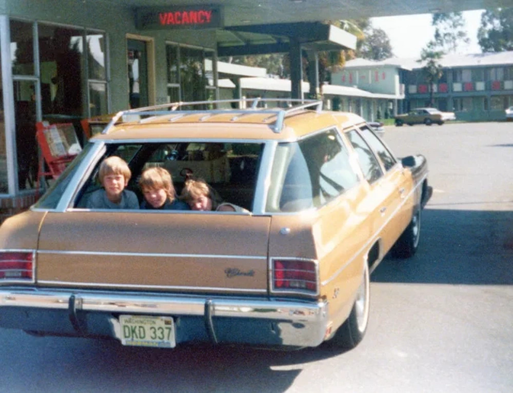Three children sit in the back of a brown station wagon parked outside a motel with a “Vacancy” sign. The car’s tailgate window is open, and buildings are visible in the background.