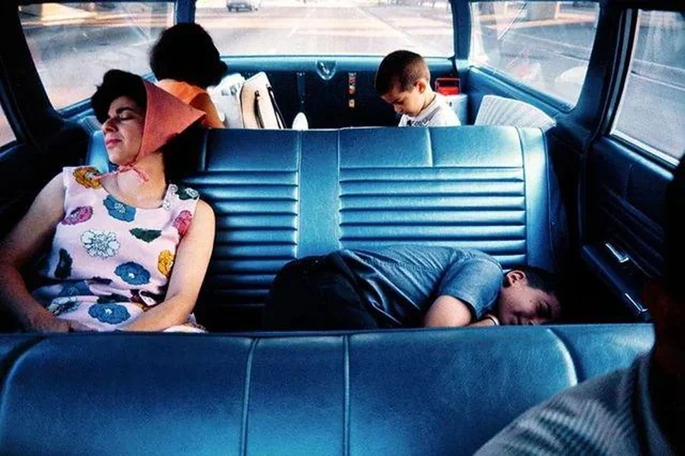 A woman rests on a car seat, a boy sleeps beside her, and two children sit in the back facing away. Sunlight streams through the car windows.