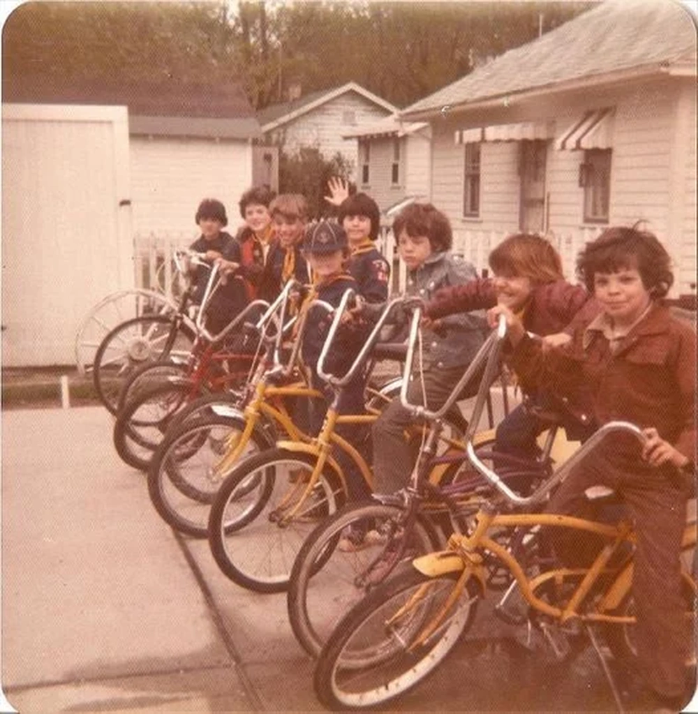 Eight children pose with their bikes in a driveway, smiling and waving. The bikes have tall handlebars. There are houses and sheds in the background on a cloudy day. The photo has a vintage, faded look.