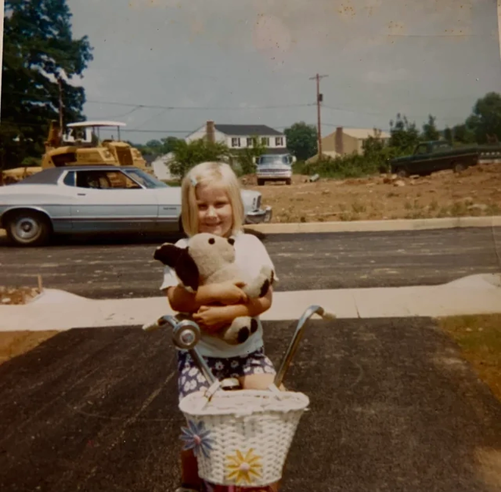 A young child with blonde hair stands on a driveway, holding a stuffed dog and a bike with a white basket. A vintage car, construction vehicles, and houses are visible in the background.