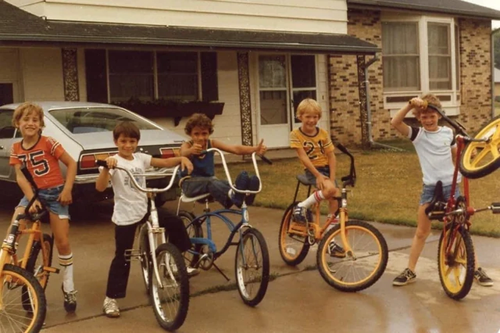 Five kids in shorts and T-shirts pose on bikes in a driveway in front of a house. One child holds a yellow bike upright. The scene looks like a summer day in the late 1970s or early 1980s.
