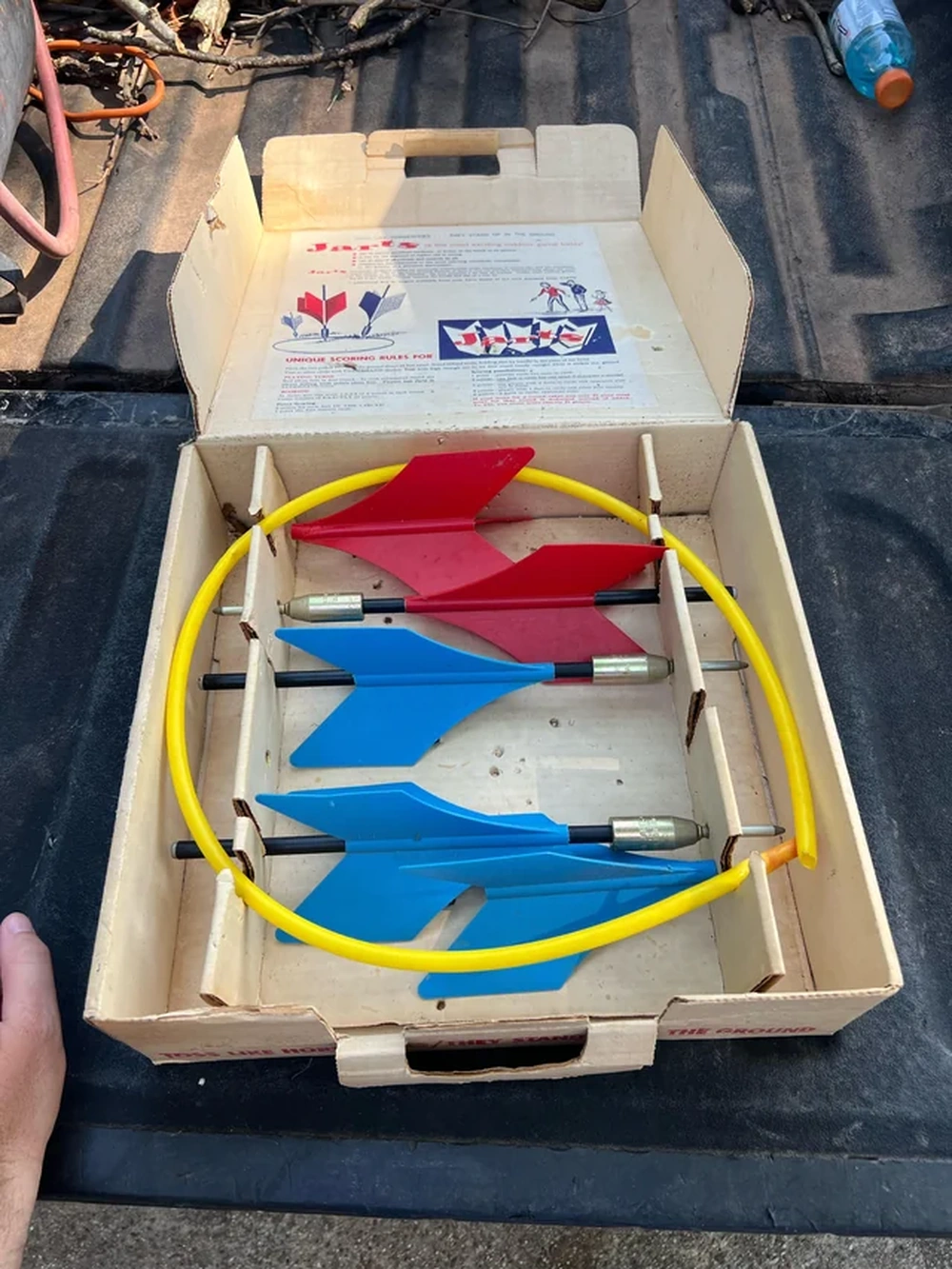 Open box containing vintage lawn darts with two red and two blue darts and a yellow plastic ring, displayed on a dark surface. Instructions are printed inside the lid.