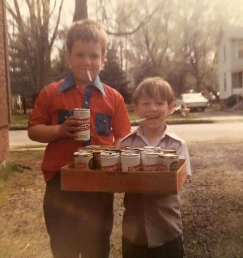 Two young boys stand outside holding a box of canned drinks, each with a straw in their mouth. The background shows trees, a car, and a white house on a sunny day.