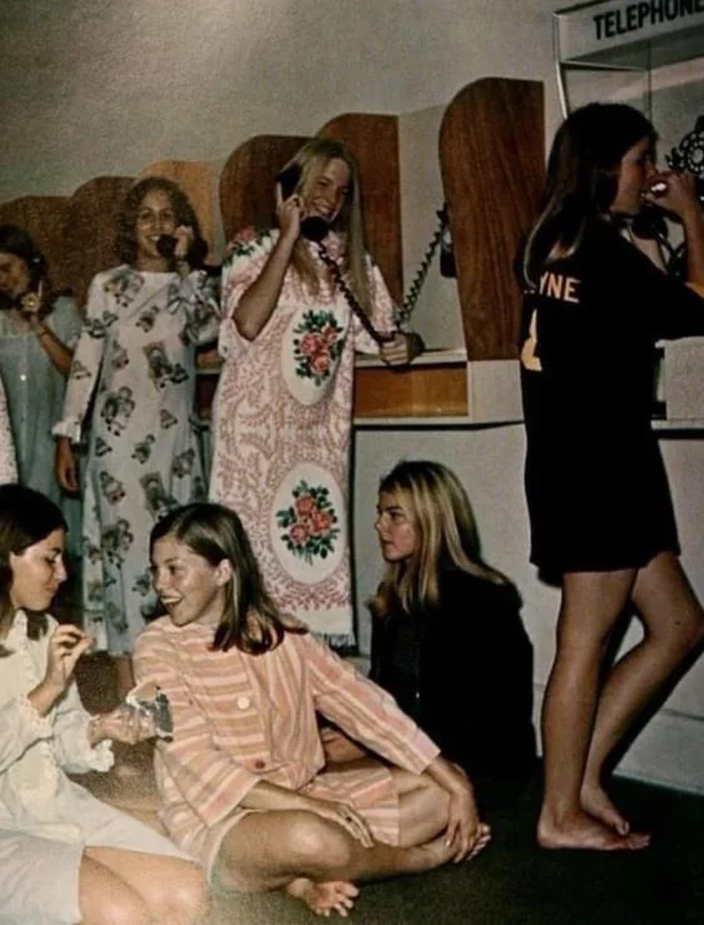 Six young women in pajamas and nightwear use rotary payphones, while two others sit on the floor chatting. The scene appears to be from the 1970s, set in what looks like a phone booth area, possibly in a dormitory.
