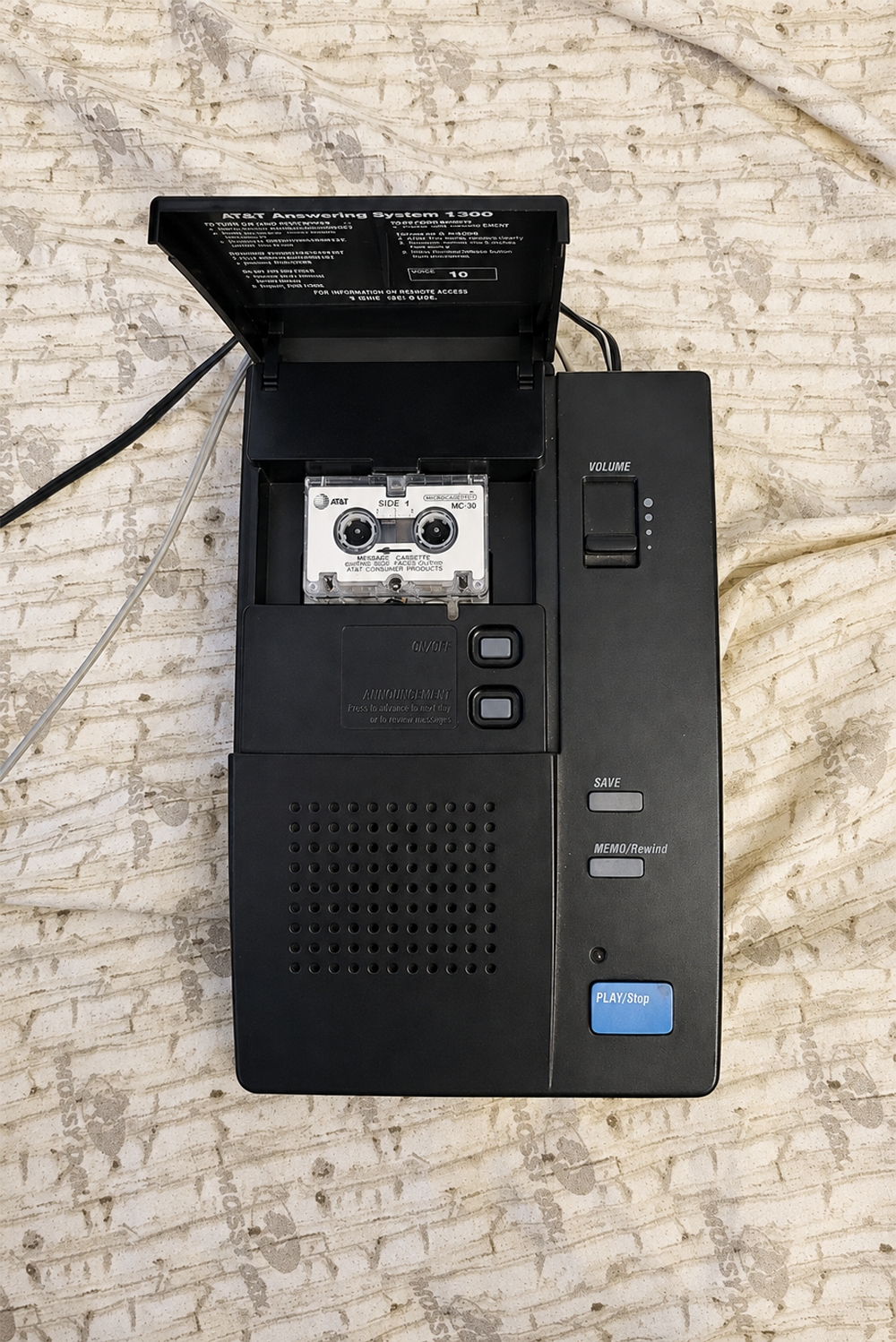 A black answering machine with an open tape deck displaying a cassette tape, placed on a beige patterned surface. The device has buttons, a speaker, and a volume slider.