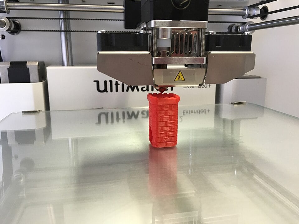 A 3D printer in the process of printing a small, red, cylindrical object on a glass platform. The printer’s nozzle is visible above the object, and the brand “Ultimaker” is seen in the background.