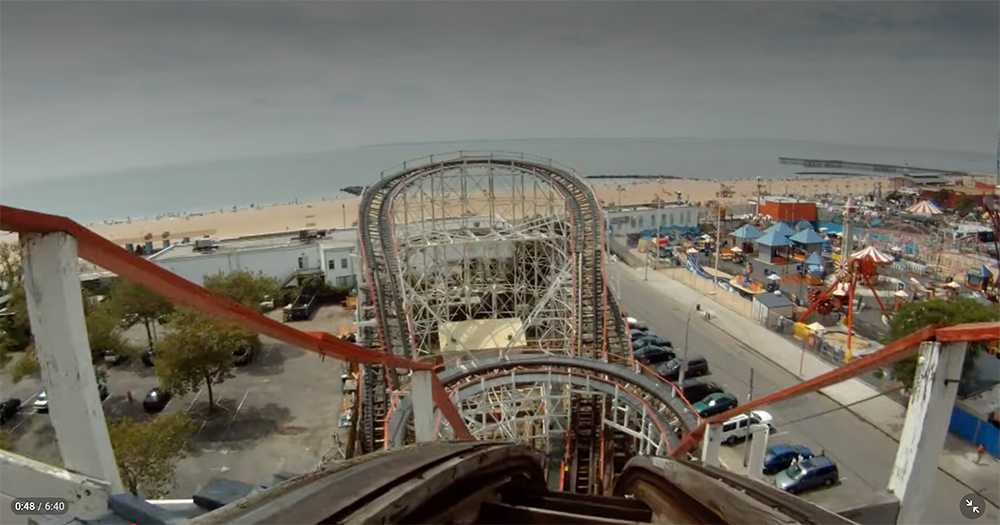 View from the top of a wooden roller coaster overlooking tracks, beach, parked cars, and an amusement park with colorful rides by the ocean under a cloudy sky.