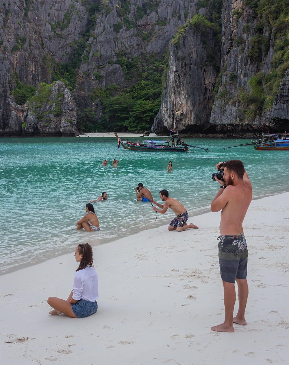 A man takes a photo of a woman sitting on a sandy beach, with people swimming in turquoise water and rocky cliffs in the background.