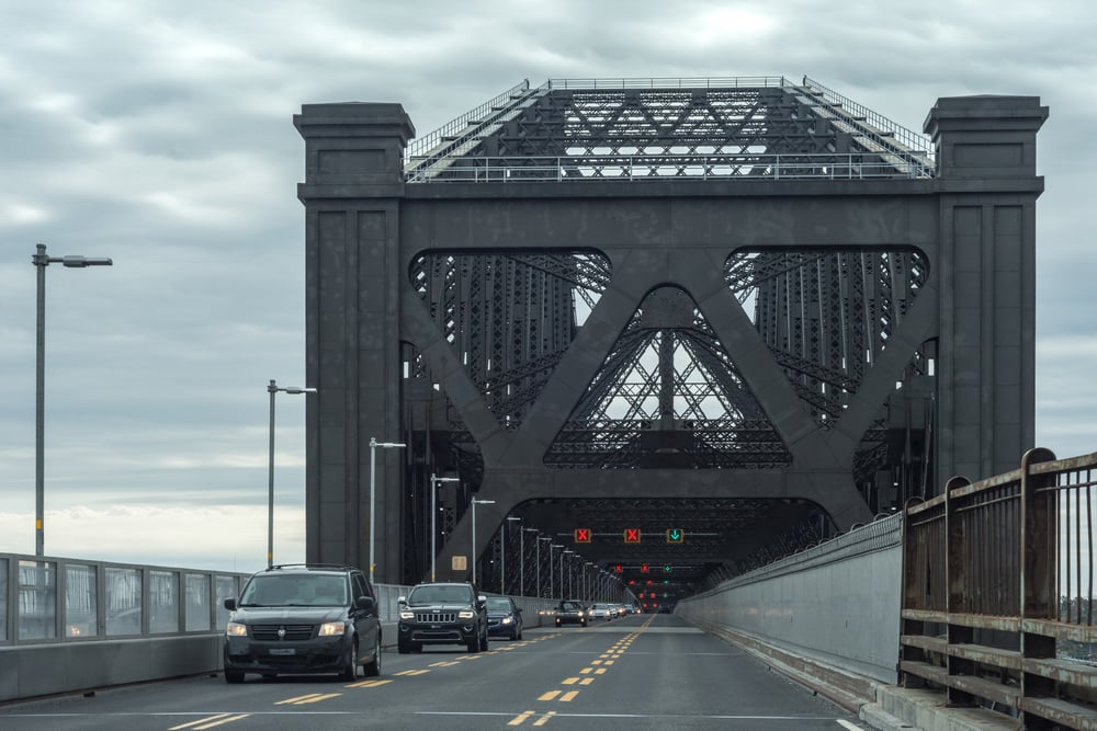 Several cars drive toward a large, dark steel bridge with intricate metalwork under a cloudy sky. Traffic lights hang above lanes, and fencing lines both sides of the road.