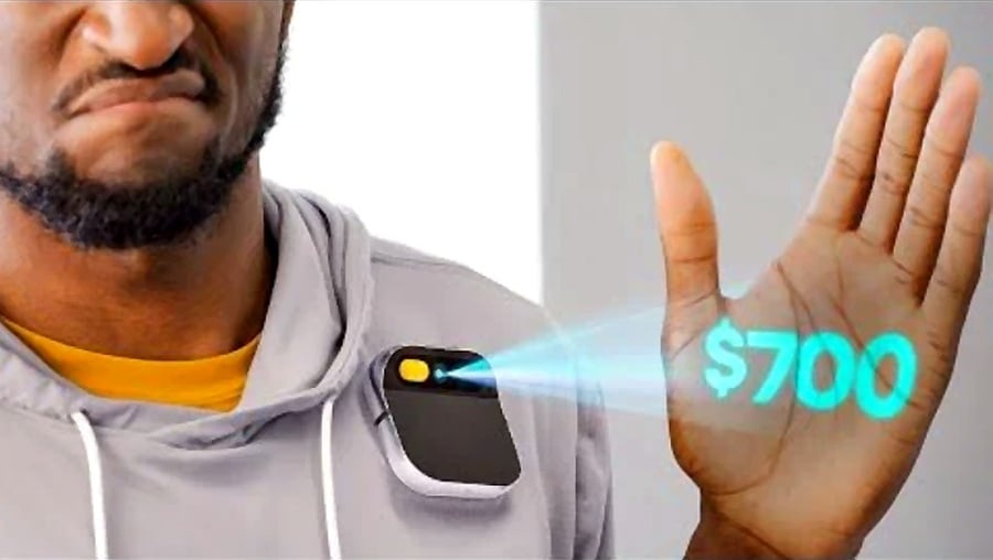 A person in a gray hoodie wears a device on their chest that projects “$700” onto their raised hand. The person appears to be frowning.