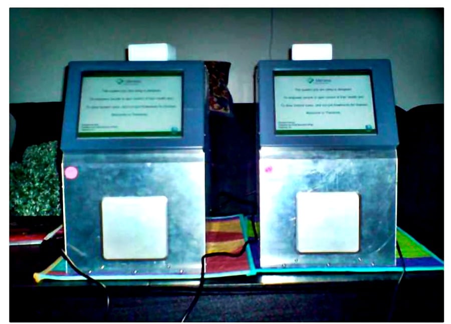 Two electronic voting machines with screens and metallic panels sit side by side on a table, each displaying text. Both have cords attached and a visible red button on the front. The background is dimly lit.