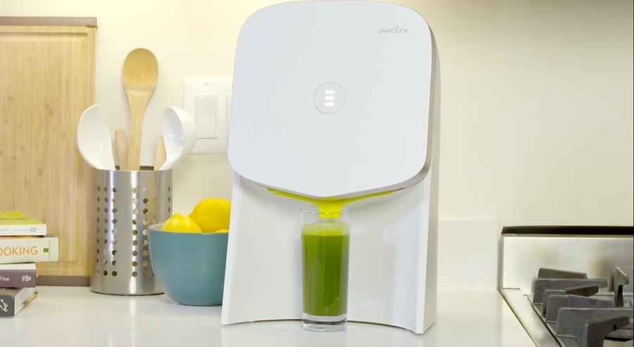 A white Juicero juicer dispenses green juice into a glass on a kitchen counter, with a bowl of lemons, utensils, and a stove nearby.