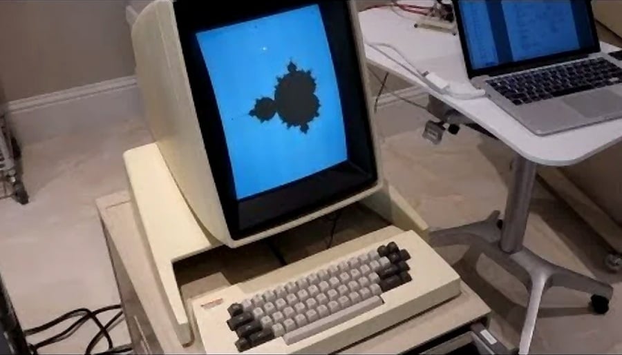 An old vintage computer displays a fractal image on its screen, with a modern laptop on a nearby desk. The computer has a beige case and a built-in keyboard with dark and light keys.