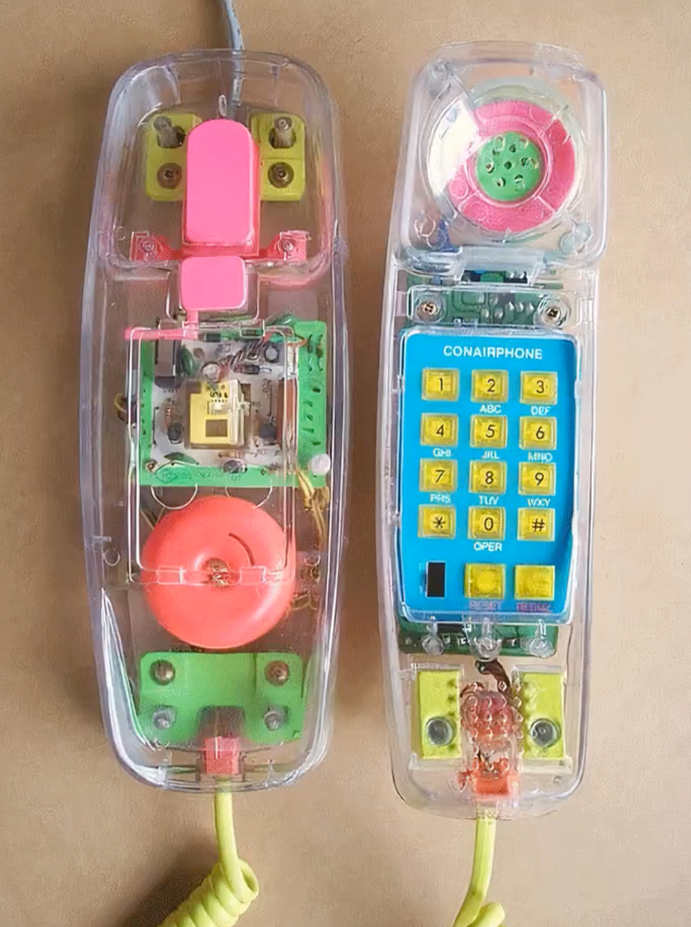 A clear plastic corded telephone with visible internal parts, pink and green components, a yellow spiral cord, and a blue number pad labeled “CONAIRPHONE,” shown with both handset and base open.