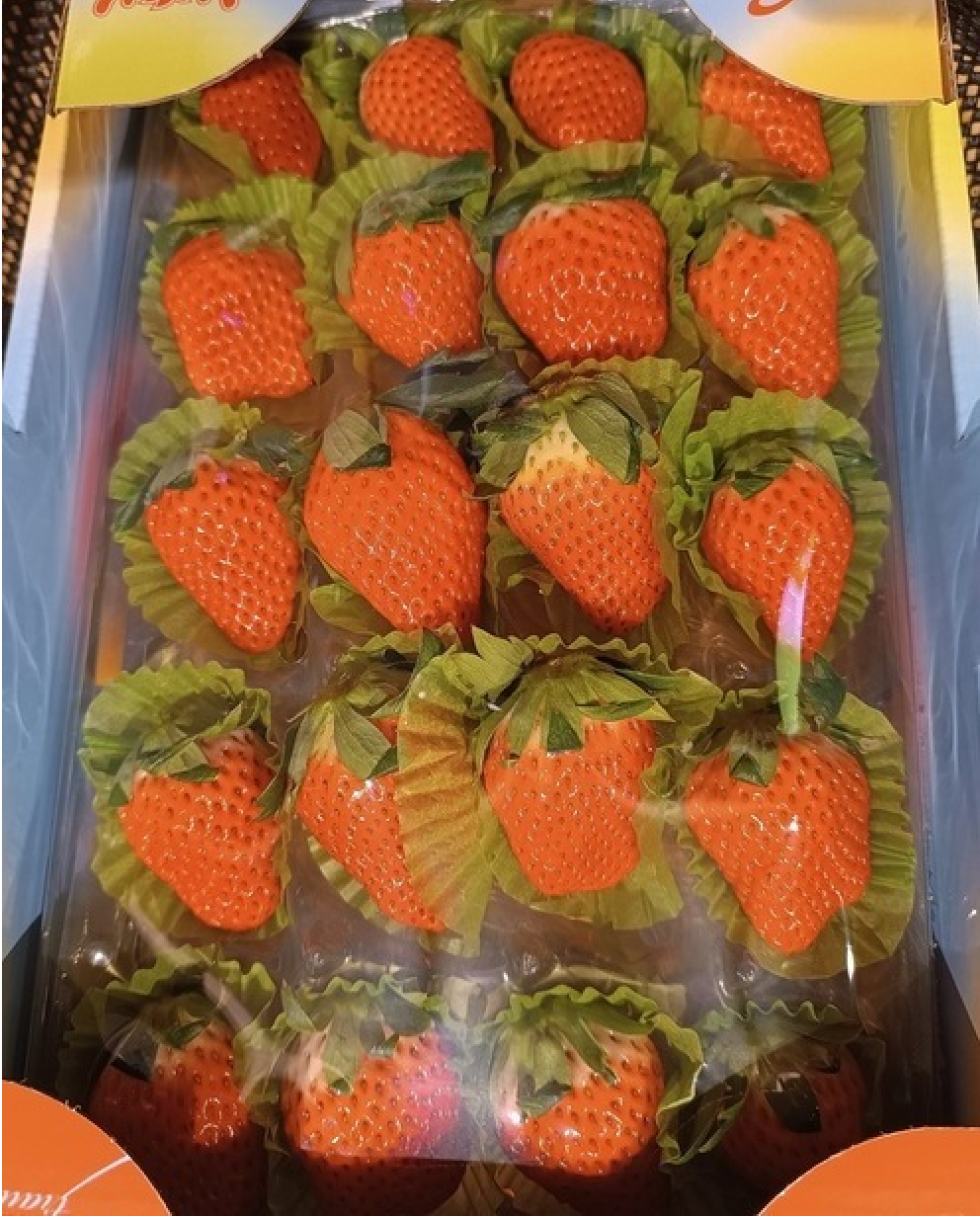 A box of neatly arranged bright red strawberries, each resting in a green paper cup, viewed from above through a clear plastic cover.