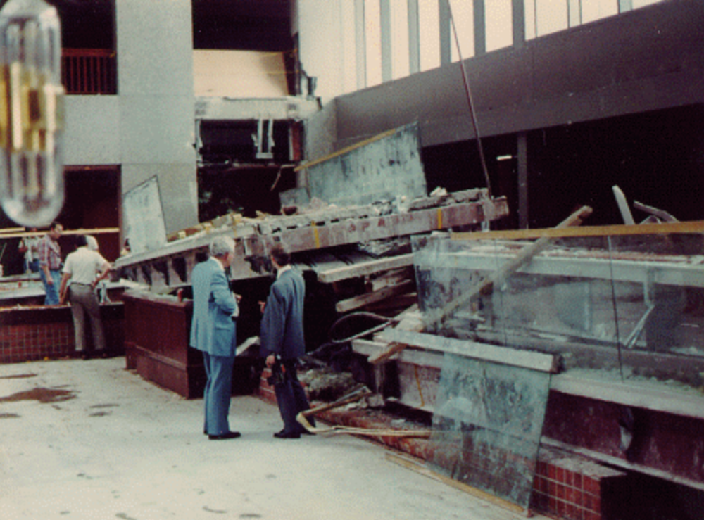Two men in suits observe a collapsed skywalk with broken glass and debris inside a large building. Other people are seen examining the damage in the background.