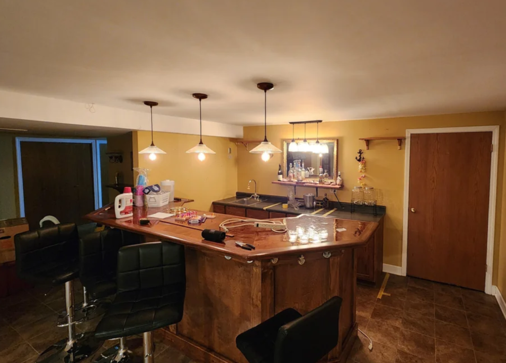 A home bar with three pendant lights, black bar stools, and a wooden counter. The counter has various items on it, and a mirror hangs on the yellow wall behind the bar.