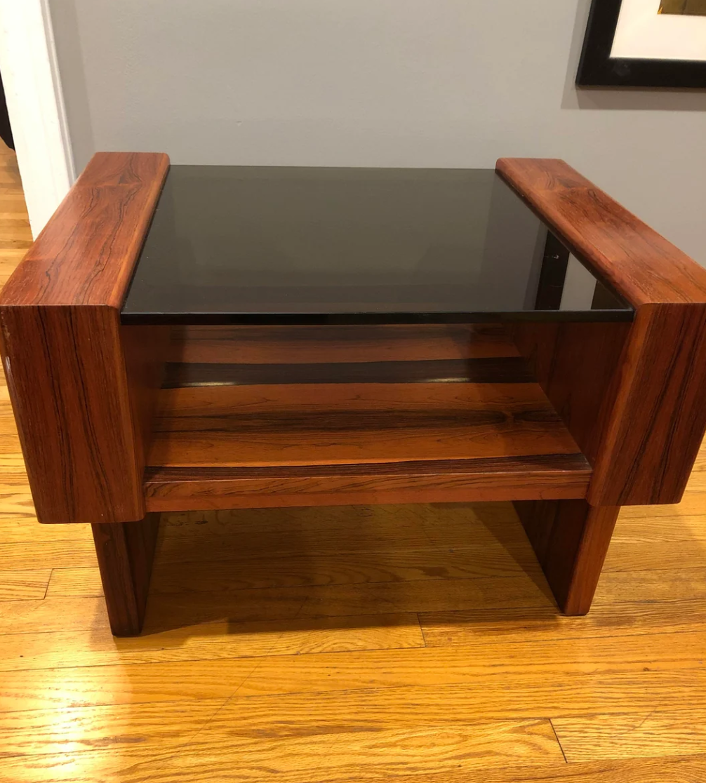 A wooden end table with a glass top and a lower wooden shelf, set on a hardwood floor against a light gray wall.