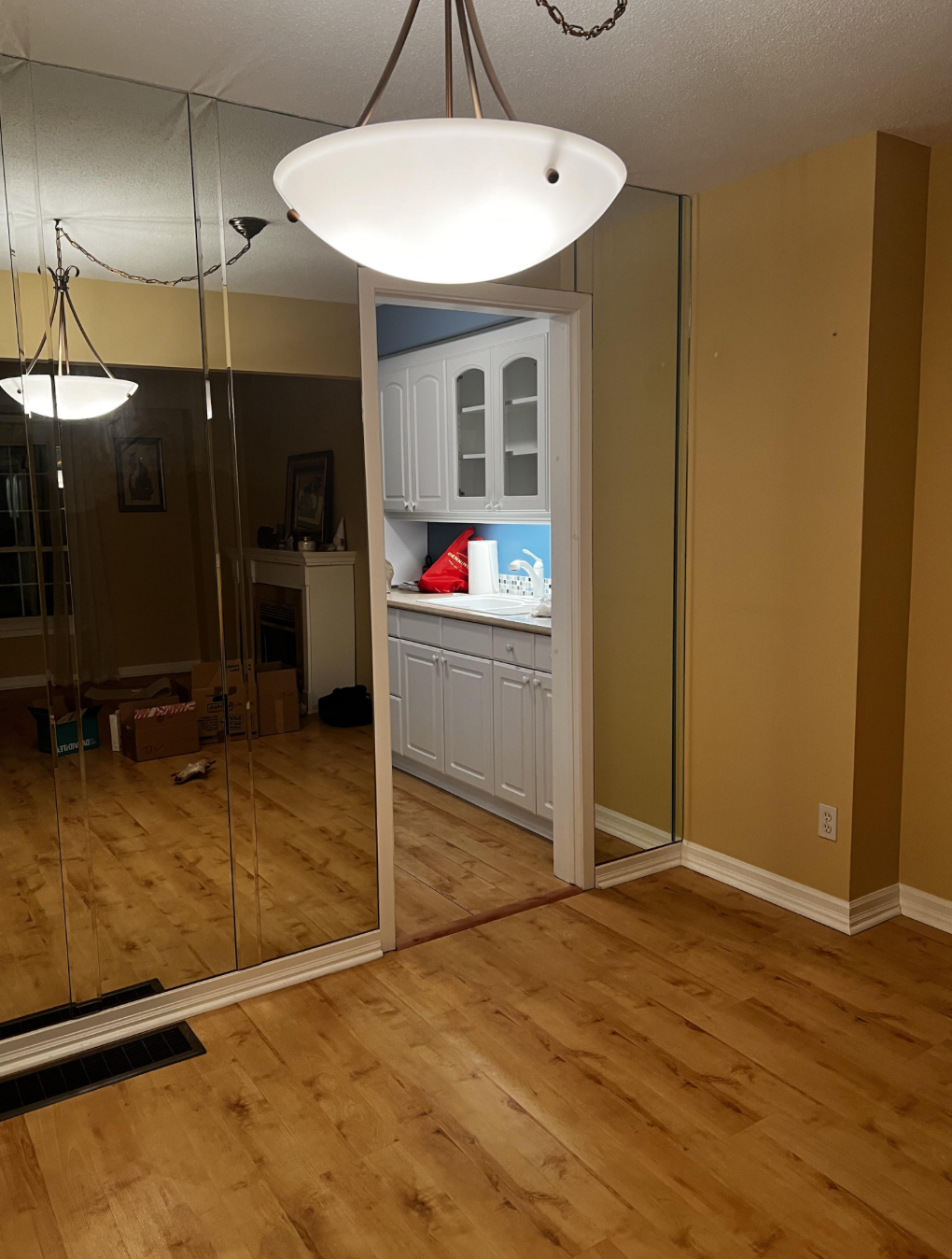 A warmly lit room with wood floors, mirrored walls, a hanging light fixture, and an open doorway showing white cabinetry and a red cushion in an adjacent room.