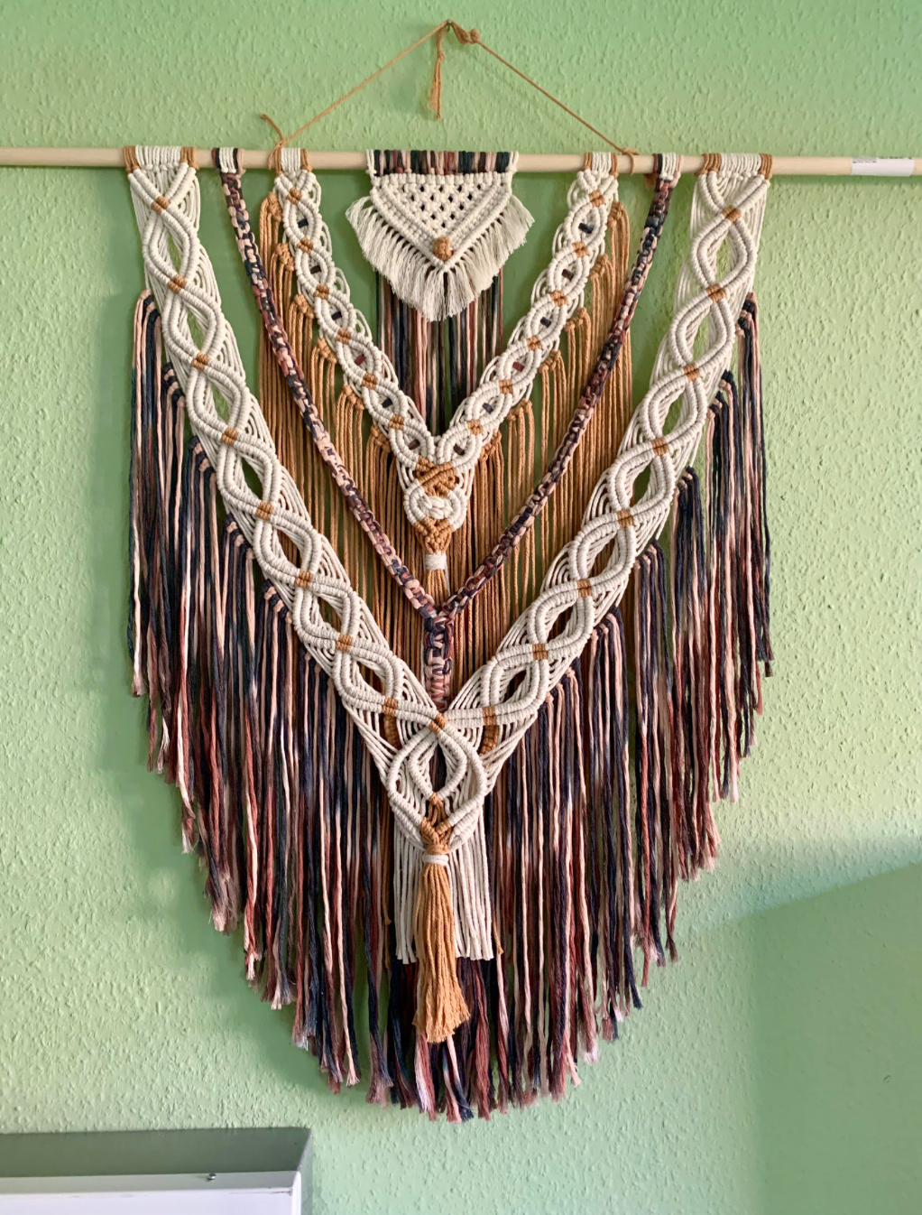 Large, intricate macramé wall hanging with white, brown, and dark blue cord patterns, featuring layered loops and fringe, displayed on a light green wall.