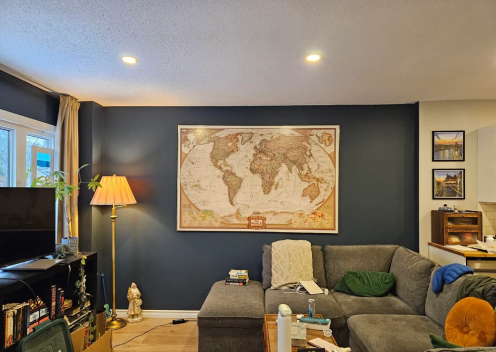 A cozy living room with a gray sectional sofa, a world map on a dark accent wall, a floor lamp, bookshelf, and decorative items. Warm lighting creates a welcoming atmosphere.