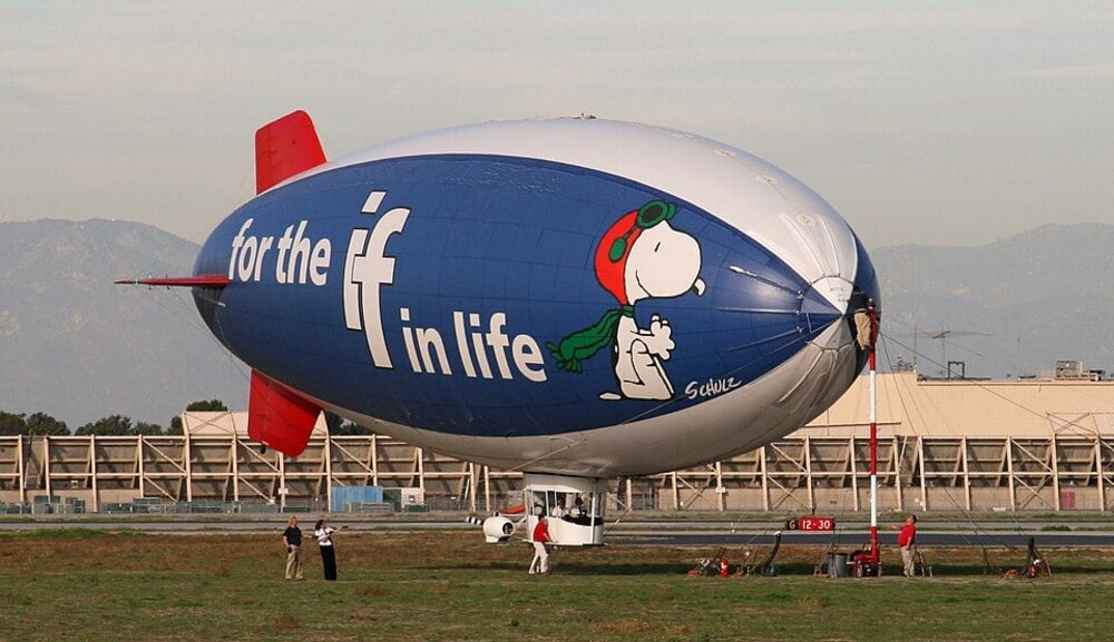 A blue and white blimp with Snoopy and the words “for the ‘if’ in life” is parked on grass, with several people standing nearby. Industrial buildings and mountains are in the background.
