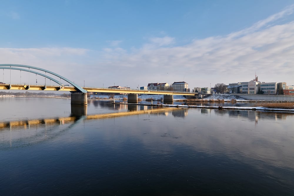 A bridge crosses a calm river under a blue sky. Buildings and snow line the far shore, and the bridge’s reflection is visible in the water.