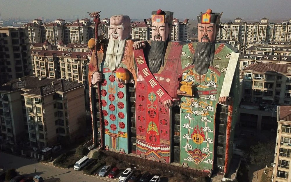 Three tall buildings painted as colorful Chinese deities stand among apartment complexes, with intricate designs and details depicting traditional robes, faces, and accessories under a clear sky.