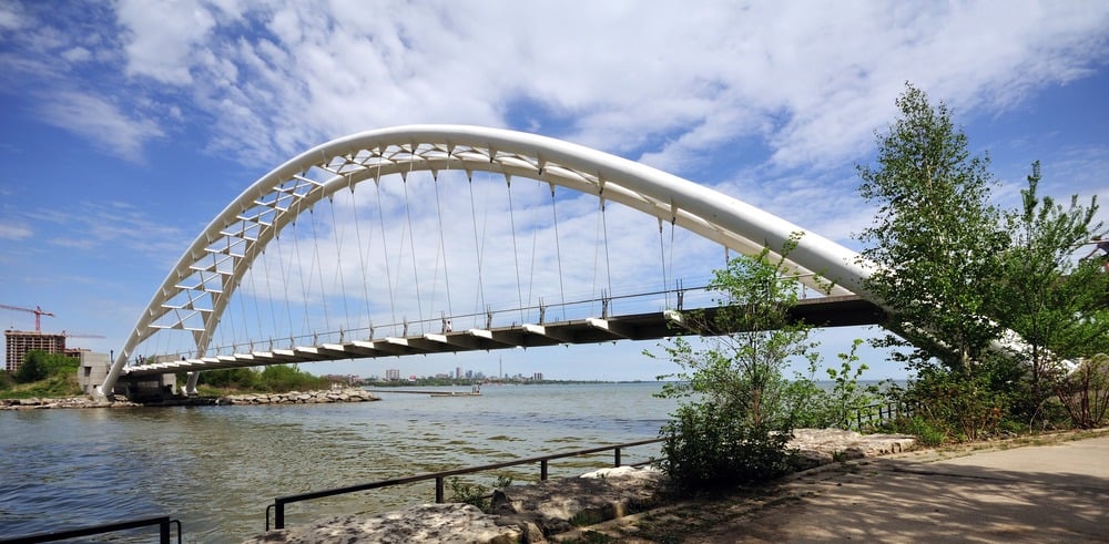 A white arched pedestrian bridge crosses over a river with a cityscape visible in the distance, trees on the right, and a partly cloudy sky overhead.