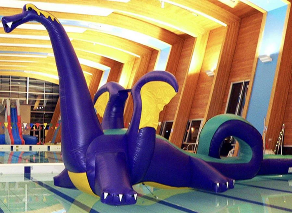 A large inflatable dragon with purple and yellow colors floats in an indoor swimming pool under wooden ceiling arches.