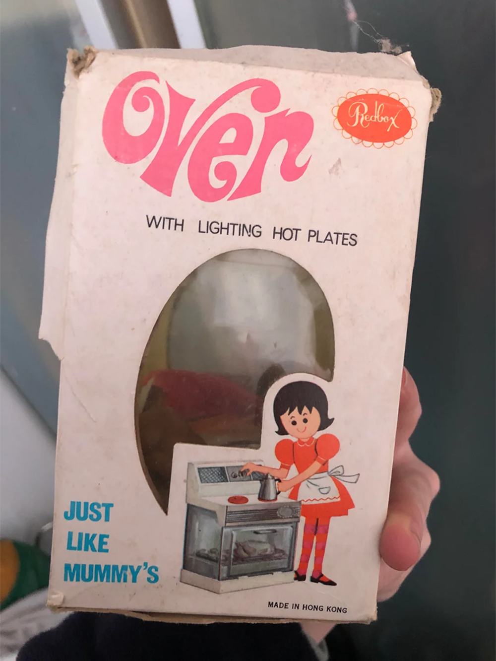 A hand holds a vintage toy oven box showing a girl illustration, an oven toy, and text reading “Oven with lighting hot plates,” “Just like mummy’s,” and “Peechox.” The box is worn and slightly torn.