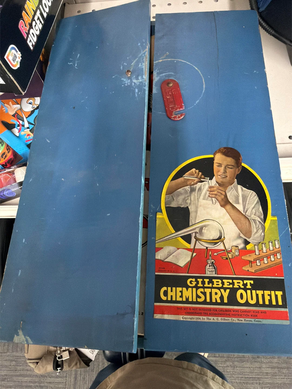 A vintage blue metal box labeled “Gilbert Chemistry Outfit” with an illustration of a boy doing chemistry experiments, partially open on a store shelf.