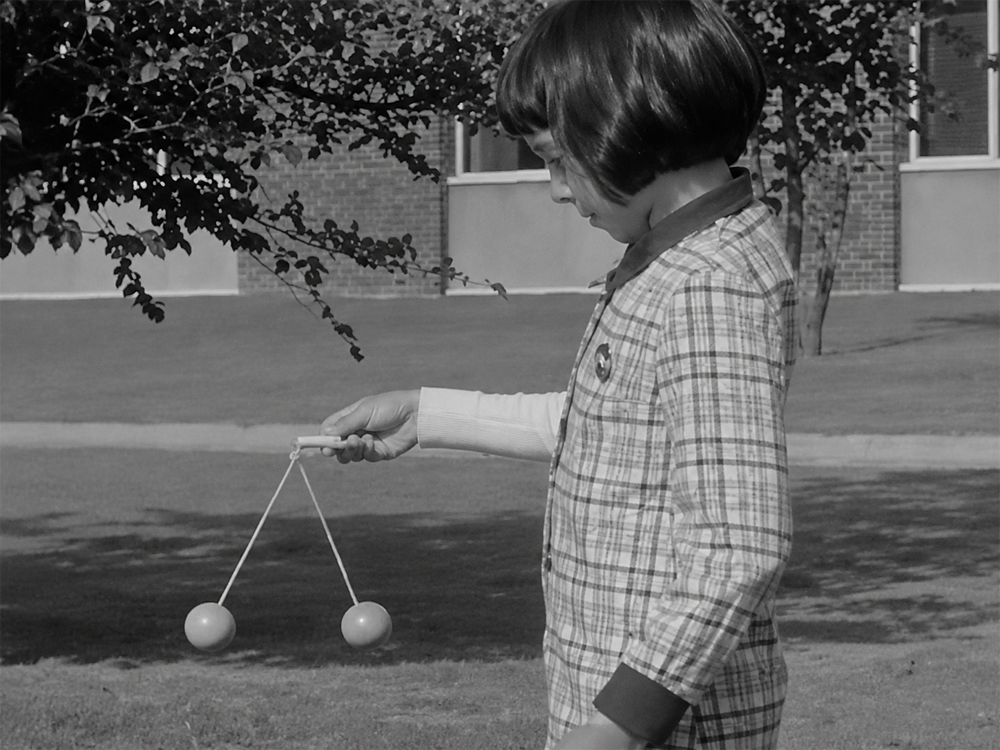 A child in plaid clothing holds a toy with two balls connected by strings outside on a grassy area, near a tree and a brick building.