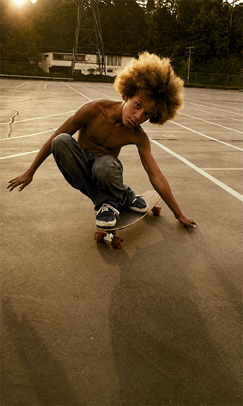 A shirtless young person with an afro skateboard on an empty outdoor court at sunset, crouching low with one hand on the ground.