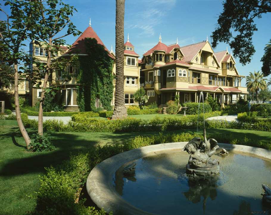 A large Victorian mansion with red roofs and many windows, surrounded by lush gardens and a circular fountain featuring a fish sculpture in the foreground under a clear blue sky.