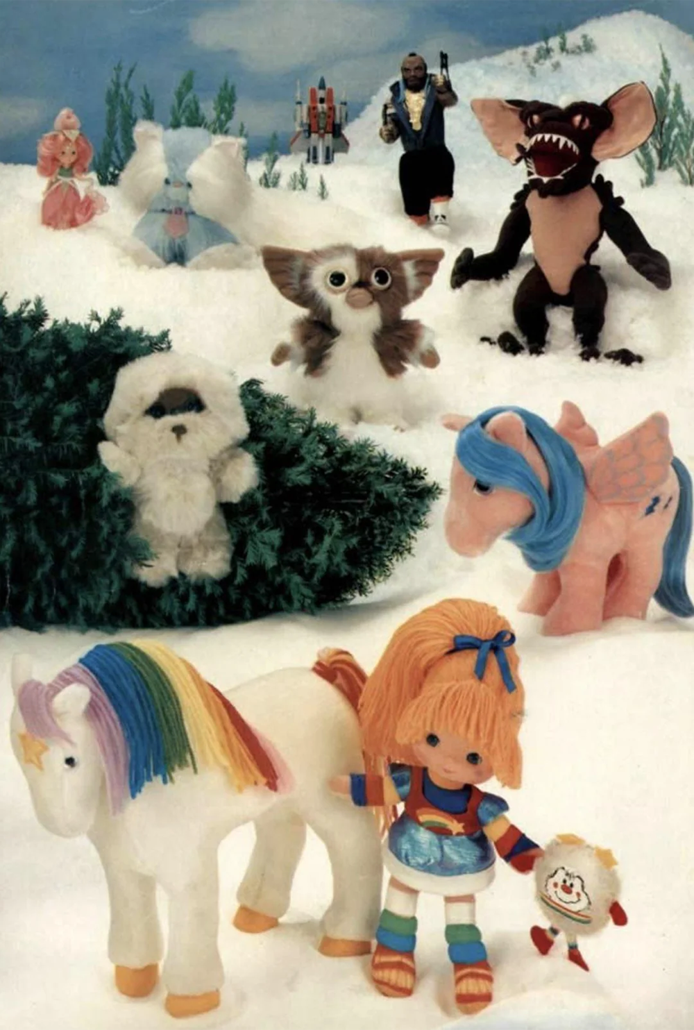 A group of 1980s toys including a rainbow unicorn, blue pony, doll, plush animals, and Gremlins figures are arranged in a snowy, wintry scene with small bushes and snow-covered ground.